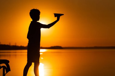 Küçük çocuk yazın güzel turuncu gün batımında nehrin kenarında kağıt bir uçağı elinde tutuyor. Gölün kenarında duran origami uçaklı bir çocuğun silueti.