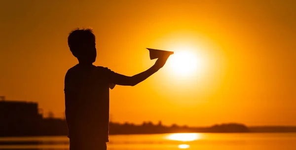 Silueta negra de un niño de pie sobre fondo anaranjado del atardecer del verano. sosteniendo un pequeño plano de papel en las manos. Fotografía horizontal . — Foto de Stock