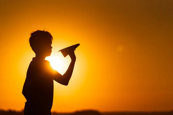 El niño sostiene un avión de papel en la mano al atardecer. Un niño levantó la mano al cielo y juega con el origami por la noche en el lago. Silueta — Foto de Stock