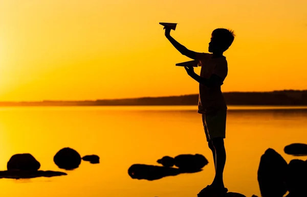 Teenie-Junge betrachtet Papierflugzeug in der Hand bei orangefarbenem Sonnenuntergang. eine Silhouette eines Kindes mit Origami-Flugzeug gegen das Wasser. Selektiver Fokus — Stockfoto