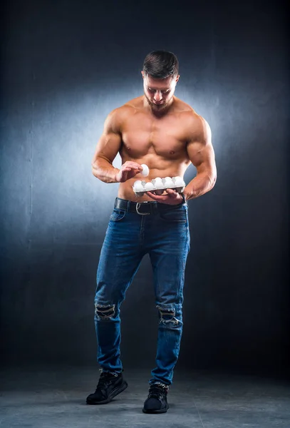 Bodybuilder joven deportista está sosteniendo un embalaje con huevos, proteínas de alimentos saludables, fondo gris. Mirando hacia abajo a un huevo. Torso desnudo, concepto de vida saludable . —  Fotos de Stock