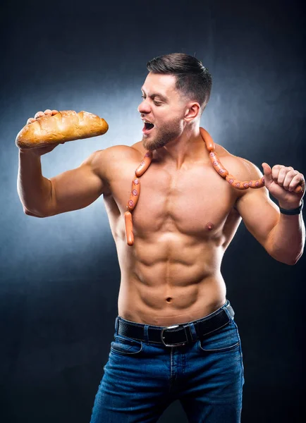 Pleased muscular guy with beautiful naked torso holding a loaf of bread and sausages on his neck. Ready to bite. Harmful food. Lifestyle concept