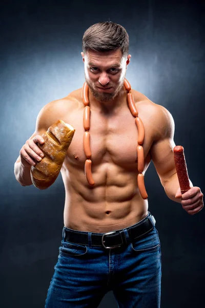 man athlete with naked torso in a black background holds bread in one hand and smoked sausage in the other hand with sausages on a neck. lifestyle concept.