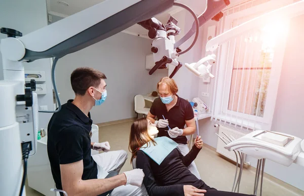 两名牙医检查病人的牙齿以进一步治疗。带有显微镜的现代口腔科柜. — 图库照片