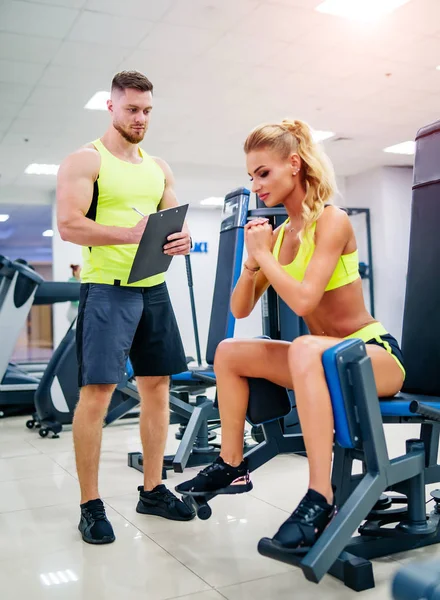 Schöne junge Frau mit ihrem Personal Trainer im Fitnessstudio diskutieren ihre Fortschritte auf einem Klemmbrett, das der Mann hält. Gesunder Lebensstil — Stockfoto