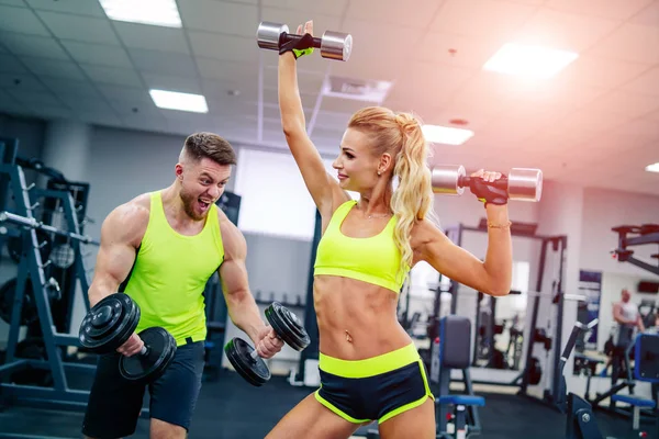 Portrait of a pretty blonde laughing and having fun with boyfriend while working out together in a gym. Hands up with dumbbell. Healthy life