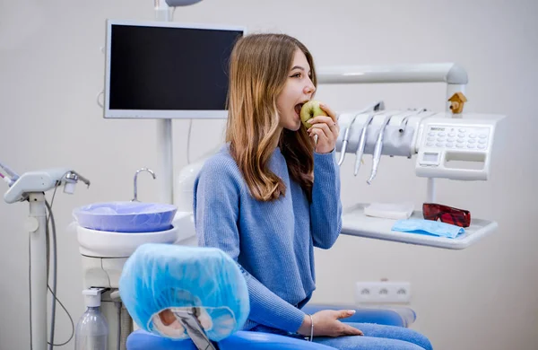 Szczęśliwa dziewczyna siedzi w fotelu dentystycznym i jeść świeże jabłko po udanym leczeniu stomatologicznym. Widok z boku. Tło gabinetu stomatologicznego. — Zdjęcie stockowe