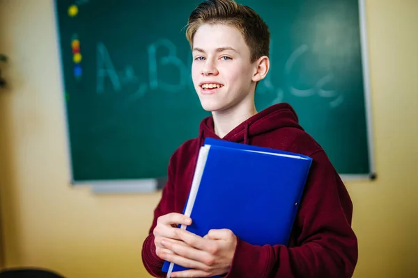 Estudante da escola higer está em pé em hoodie escuro perto do quadro negro verde na sala de aula. Adolescente Menino com caderno azul nas mãos está olhando para a câmera. Encerramento. — Fotografia de Stock