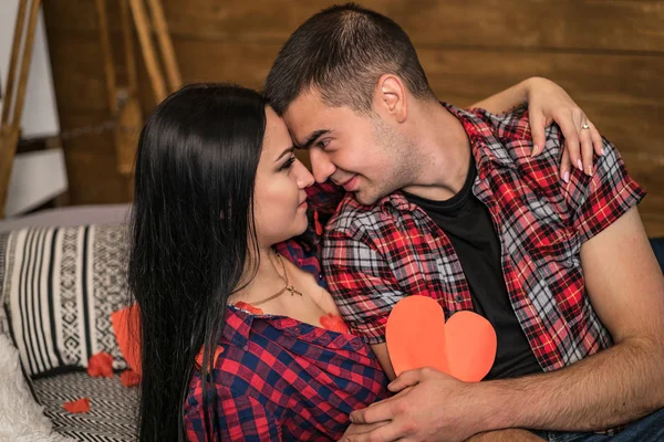 El amor real. Hermosa pareja jovencita que se acaricia y sonríe mientras se sienta en el estudio con el corazón de papel mirando el uno al otro. Concepto de historia de amor. — Foto de Stock