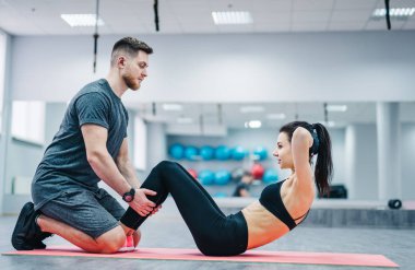 Spor salonundaki minderde koçunun yardımıyla karın egzersizi yapan genç bir kadın. Kas çalıştırıcı çekici dişinin karın kaslarını çalıştırmasına yardım eder
