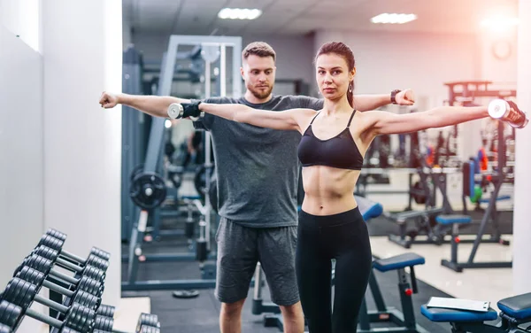 Lächelnde Frau in schwarzem Top und Leggins mit Hanteln vor Trainerin im Fitnessstudio. Männlicher Trainer und schöne Frau beim Heben von Metallhanteln. — Stockfoto