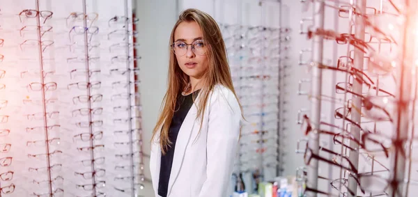 Female client, optician is standing with set of glasses in background in optical shop. Stand with spectacles. Eyesight correction. Girl wearing glasses. Portrait of a woman in correcting glasses. — 스톡 사진