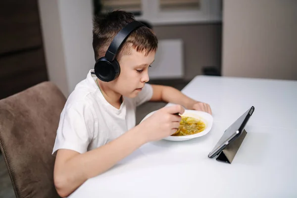 Niño comiendo y jugando en la tableta con auriculares en casa —  Fotos de Stock