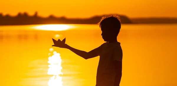 El barco de papel está en manos de un niño. Hermosa puesta de sol de verano. Origami — Foto de Stock