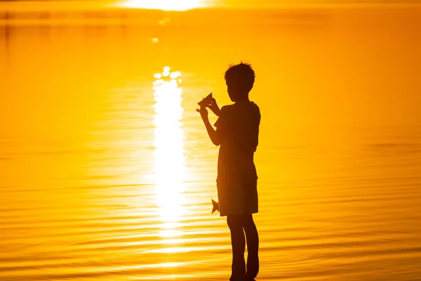 Rapaz, lança um navio de papel na água. Belo pôr-do-sol de verão. Barco de papel. Origami . — Fotografia de Stock