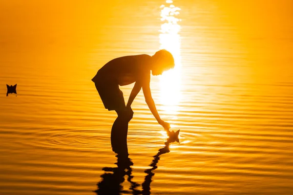 El niño lanza barcos de papel en la playa en verano. Hermoso atardecer. Origami. Río. Lago. . — Foto de Stock