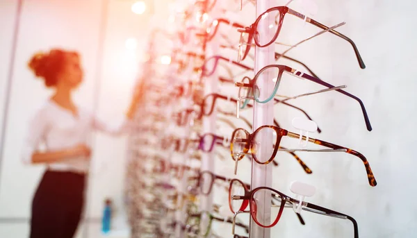 Fila de gafas en una óptica. Tienda de anteojos. Soporte con gafas en la tienda de óptica. La mujer elige los anteojos. Corrección de la vista. — Foto de Stock