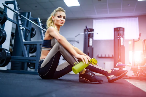 Mujer fitness sosteniendo la botella de agua mientras está sentada y descansando en el suelo en el gimnasio. Concepto de vida saludable — Foto de Stock