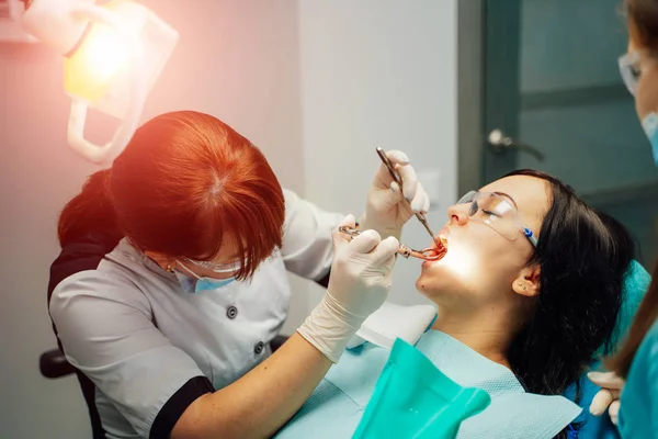 Stomatologin und Assistentin in Masken und Schutzbrille bei der Arbeit mit einer Patientin in der Zahnarztpraxis. Patientin sitzt in Klinik im Zahnarztstuhl — Stockfoto