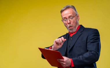 Kıdemli işadamı, sarı arka planda dosyasıyla oturuyor. Şaşkın ve komik görünüyor. İş konsepti