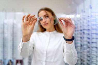 Kadın müşteri ya da gözlükçü optik dükkanında arka planda çiğ bardakla ayakta duruyor. Gözlükleri takın. Görme düzeltmesi. Gözlüklü kız kameraya poz veriyor..