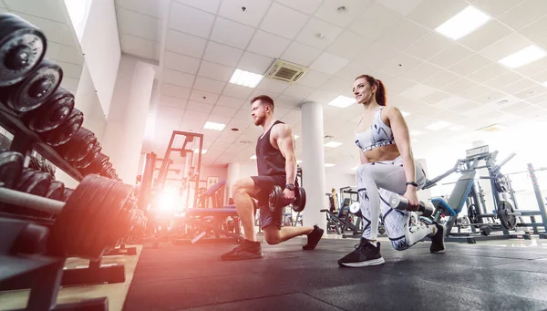 Healthy couple in sport clothes lifting dumbbells in the gym. Attractive woman and handsome man doing workout with dumbbells standing in special pose in the sports club