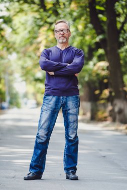 Açık havada gri saçlı orta yaşlı bir adam. Mavi kazak. Gözlük. Sakallı, kendine güvenen iş adamı. Arka plan bulanık. Çapraz eller.