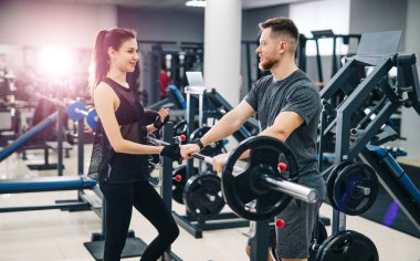 Genç, güzel esmer fitness kızı spor aletleriyle spor salonunda antrenman yapıyor. Antrenör daha iyi sonuçlar için önerilerde bulunuyor.