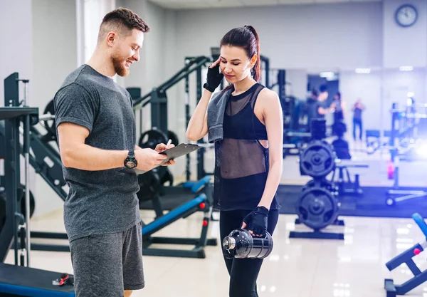 Schöne junge Frau mit ihrem Personal Trainer im Fitnessstudio diskutieren ihre Fortschritte auf einem Klemmbrett, das der Mann hält. Gesunder Lebensstil — Stockfoto
