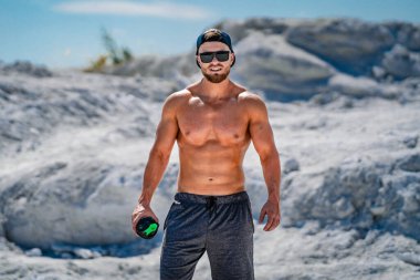 Gözlüklü ve şapkalı yakışıklı, üstsüz vücut geliştirici. Sporcu elinde bir şişe suyla kameranın önünde duruyor. Dışarıdaki fotoşop..