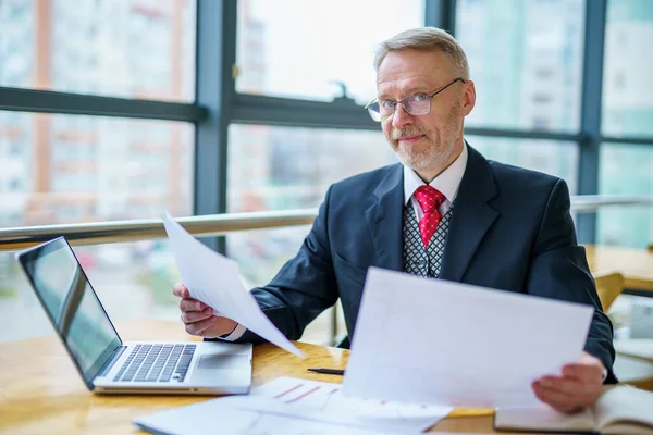 Dojrzały Biznesmen Oglądający Laptopie Drukowane Dokumenty Siedzi Przy Oknie Biurze — Zdjęcie stockowe