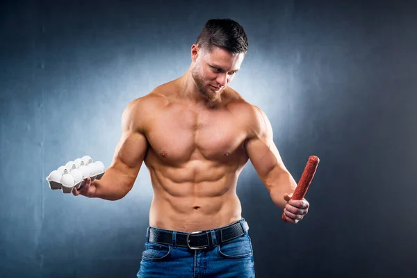 Bodybuilder sportsman holding eggs and sausage . Choosing between healthy and harmful food. Naked torso. Grey background. Closeup