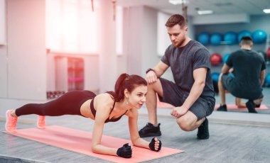 Tatar-fit kadın spor salonundaki pembe bir paspasın üzerinde dirsek dirseğiyle şınav çekiyor. Koyu renk saçlı, spor hocası olan, kalas ve dirsek kayışı takan bir kadın. Yakın plan.