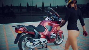 Kasklı güzel bir kadın, kırmızı bir motosikletin arkasında siyah bir kıyafetle şehir manzarasına karşı oturuyor.
