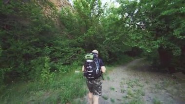 Sıcak bir yaz gününde, sırt çantalı ve tişörtlü bir turist, kayalıkların yakınındaki ormanda bir patika boyunca ilerliyor.