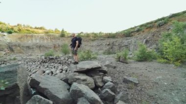 Sırt çantalı yetişkin gezgin nehir kenarındaki araziyi inceliyor ve harika bir manzaranın arka planında kıyıdaki kayalar boyunca yürüyor.