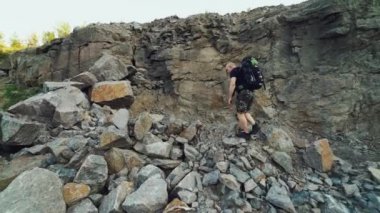 Omuzlarında sırt çantası olan siyah tişörtlü ve şortlu ekstrem turist, yazın sıcak bir havada kayalıklarda yürüyor ve büyük bir kayaya tırmanıyor.