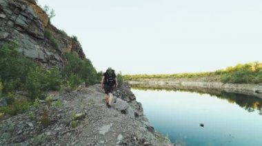 Sırtında sırt çantası olan turist, gölün kıyısında yüksek kayalıkların ve vahşi doğanın arka planında yaz günü ilerliyor.