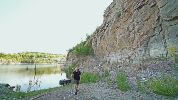 去年夏天 一位成年旅游者背着背包 提着手杖 背着高高的岩石 沿着湖岸走着 — 图库视频影像