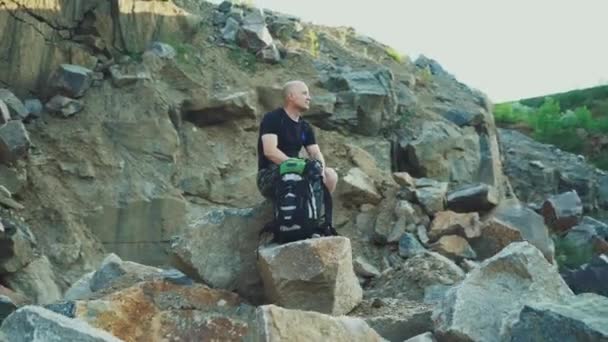 彼の隣に石の上に座って自分の周りの岩で素晴らしい自然を見ているリュックを持つ大人の観光客 — ストック動画
