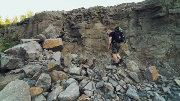 黒のTシャツと肩にリュックを背負ったショートパンツの極端な観光客が岩の上を歩き夏の暖かい天候の中で大きな石を登る — ストック動画