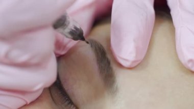 Eldivenli kozmetik uzmanı kadın kaşlarının şeklini profesyonel bir iğneyle düzeltiyor. Kalıcı kaş makyajı prosedürü.