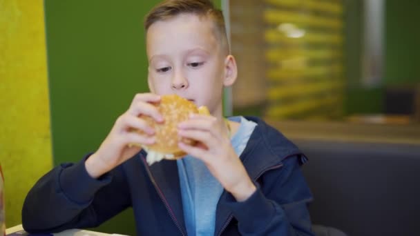 ファーストフードのカフェで男の子 ファーストフードのカフェでハンバーガーとフライドポテトを食べる 有害な栄養 — ストック動画