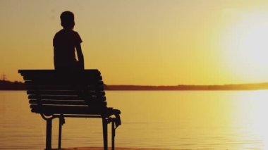 Küçük bir çocuk gün batımına ve nehrin kenarındaki bankın üzerindeki muhteşem manzaraya bakıyor. Bir adam ellerini kaldırıyor. Hafta Sonu.