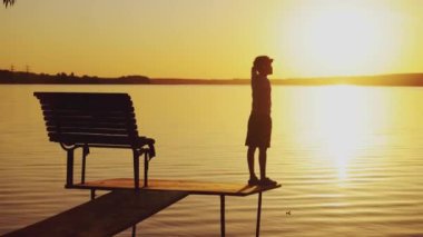 Küçük bir çocuk bankın yanındaki duvarda duruyor. Adam yazın göl kenarında gün batımında etrafa bakıyor..