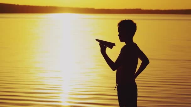 一个小男孩手里拿着一架纸飞机 在夕阳西下的河岸上与他一起在太阳能跑道上玩耍 — 图库视频影像
