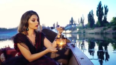 Siyah ve kırmızı elbiseli güzel bir model elinde dekoratif bir fener tutuyor ve akşamları su ve gökyüzünün arka planında gondolda poz veriyor. Venedik 'teki karnaval. Bulanık Arkaplan
