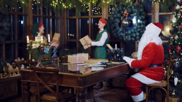 サンタクロースはタイプライターに手紙を書いていて エルフが新年の前夜に部屋でクリスマスの装飾の背景にお互いにプレゼントを投げているのを見ています — ストック動画
