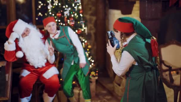 除夕之夜 一位小精灵助手正在圣诞树下给圣诞老人和小精灵拍照 并在窗边用复古相机装饰着花环 模糊的背景 — 图库视频影像