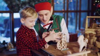Uzun kulaklı ve kırmızı şapkalı bir elf elinde bir büyüteç tutuyor ve küçük bir çocuğa arka planda neşeli bir oyuncak ve Noel Baba 'nın evinin penceresinin yanında bir Noel ağacı gösteriyor.